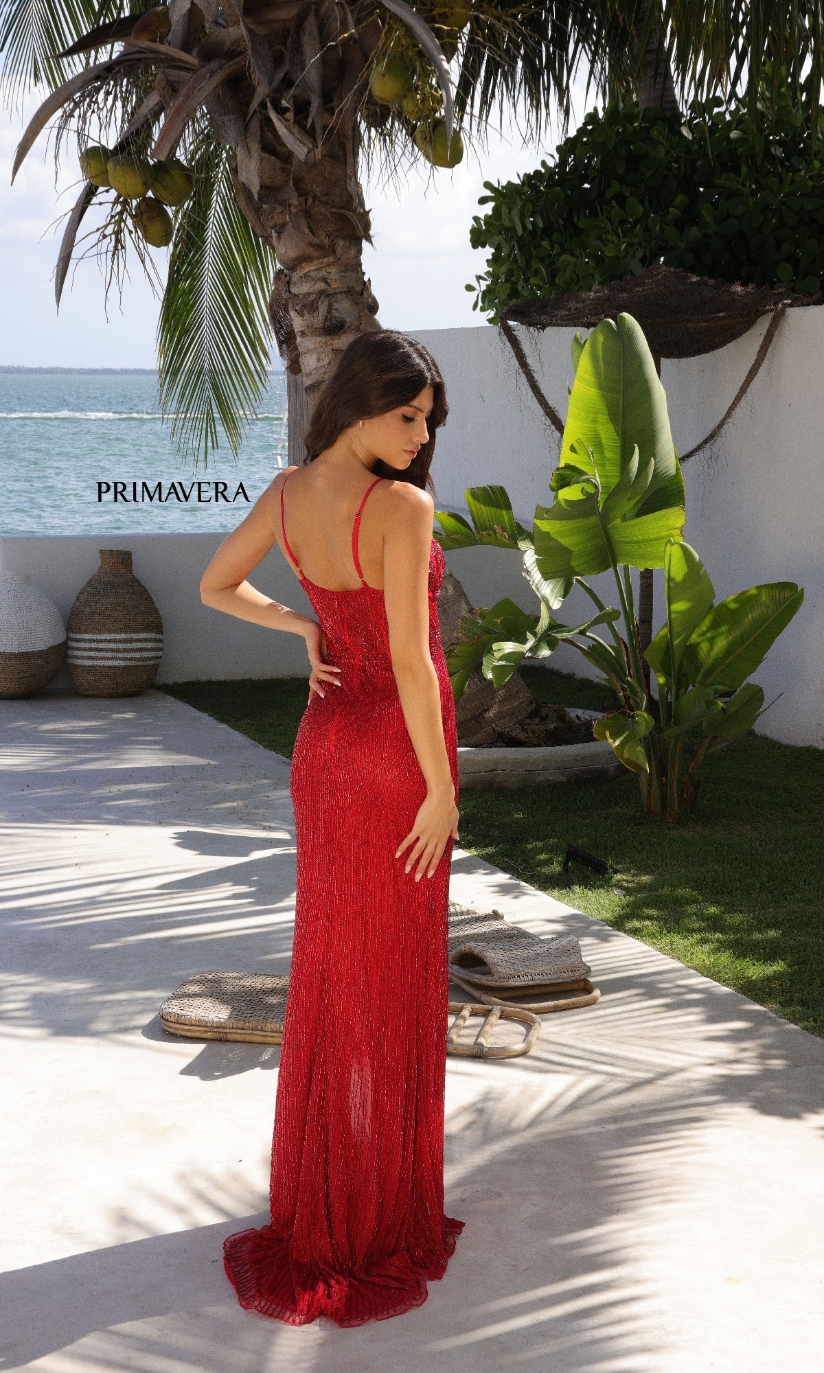 A woman stands outdoors among tropical plants, facing away while wearing the Primavera 4352 Cut-Out Long Beaded Prom Dress—a sleeveless, floor-length red gown.